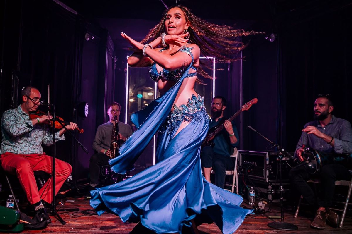 A bellydancer wearing a turquoise belly-dancing outfit is performing with long hair flying in front of a traditional ensemble of four musicians, playing the violin, tabla, saxophon,e and guitar. 
