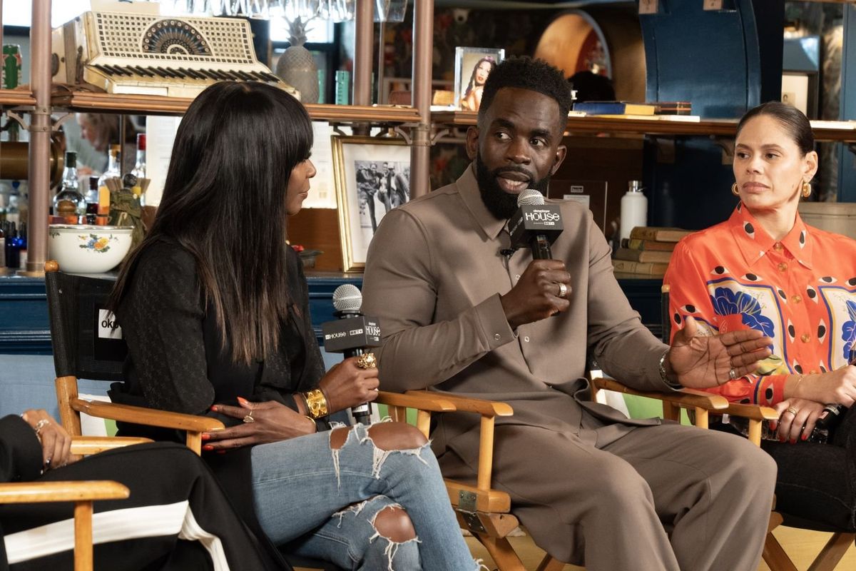 A Black man in a brown suit sits in a studio chair with four other people and speaks into a microphone while everyone pays attention.
