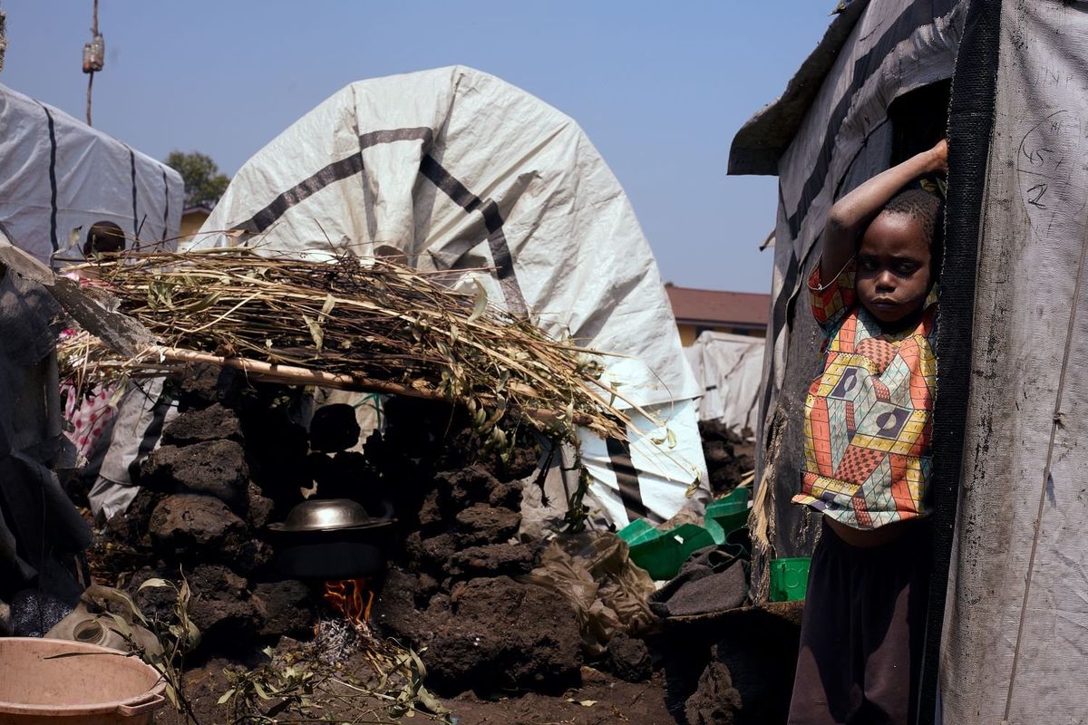 A child hangs onto a tent near a cooking pot over a fire. 