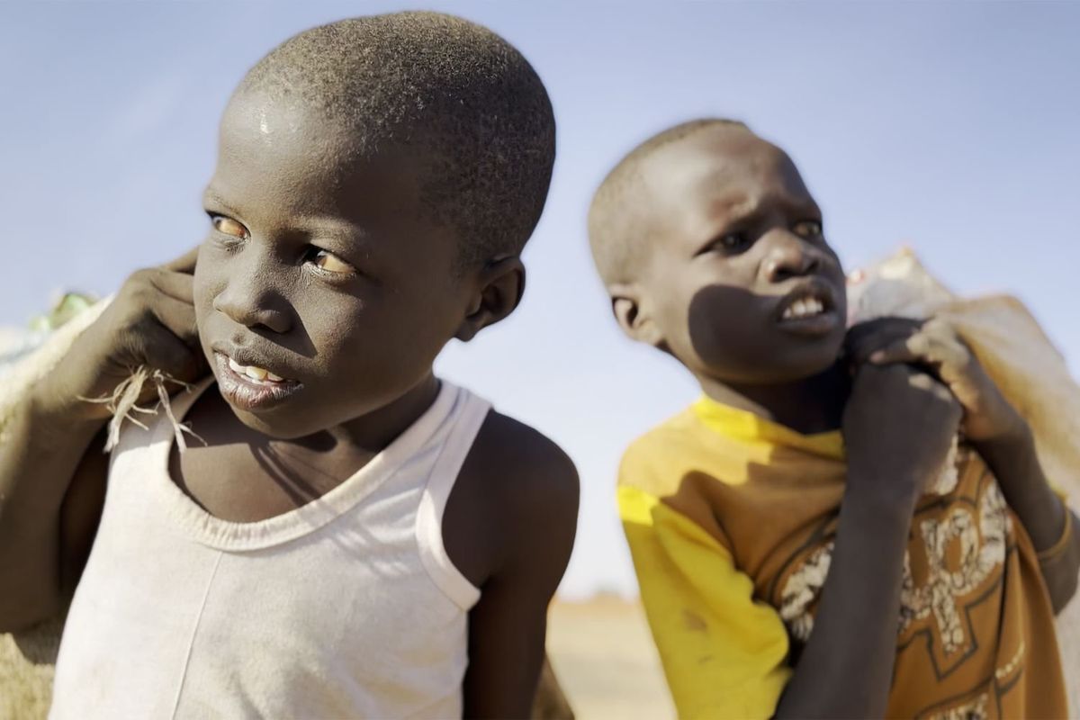 A close-up shot of two boys, one wearing a white tank top and one a yellow t-shirt, carrying plastic bottles in sacks on their backs. 