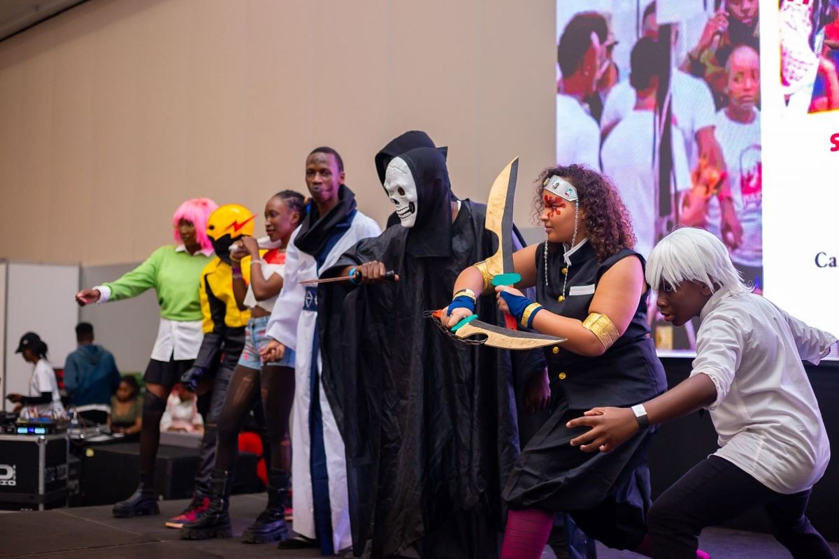 A diverse group of cosplayers in various costumes, including a character with a scythe, a character with twin blades, and others in colorful outfits, strike poses on stage at the cosplay competition during NAICCON.