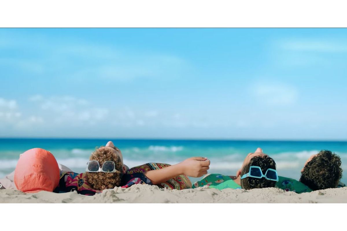 A group of four young men are lying on the beach, the turquoise Mediterranean sea in the background and only the back of their heads are showing. Two of them have