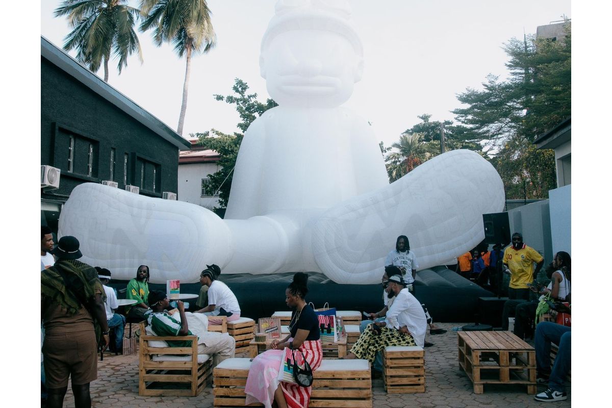 A group of people gather around wooden pallet seating in an outdoor courtyard, in front of a massive white inflatable figure with goggles, surrounded by palm trees and buildings.