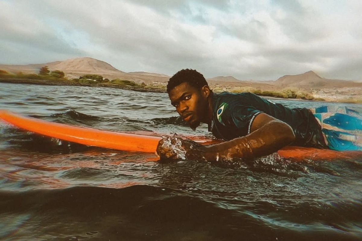 A man lies on a bright orange surfboard in the water, looking intensely toward the camera, with mountains and cloudy skies in the background.
