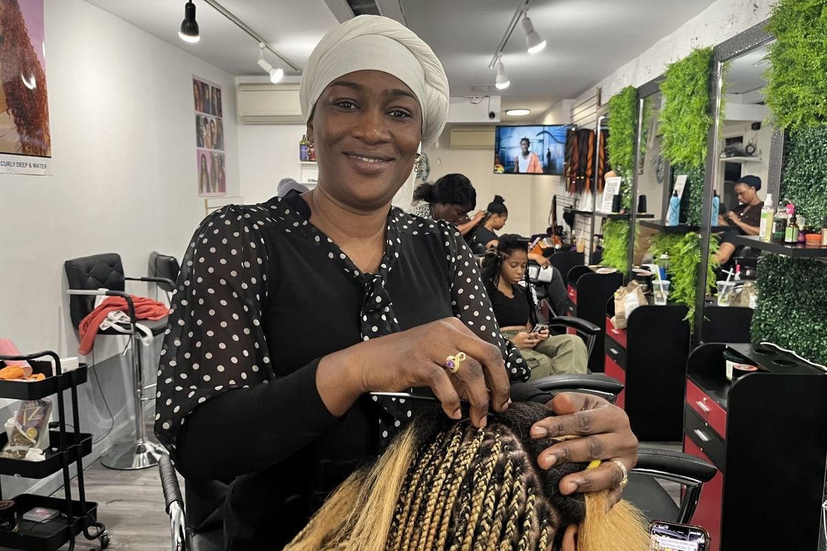 A photo of Nikiama Kisun Sira Fatimata in her salon braiding a client’s hair, and wearing a black blouse with white Polka Dot sleeves.