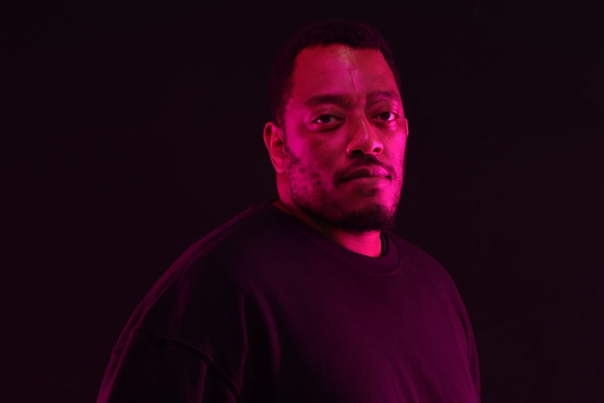 A portrait photo of Likarion Wainaina wearing a black sweatshirt and blue denim pants in a dimly lit room.