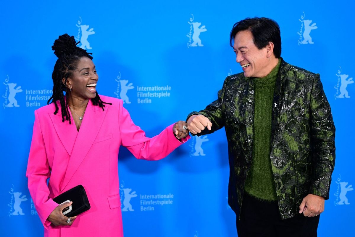 A smiling Black woman with long locks styled in a top bun, wearing a bright pink suit, fist-bumps an Asian man in a textured green suit and sweater; they stand in front of a blue backdrop for the Berlin International Film Festival, sharing a joyful moment.