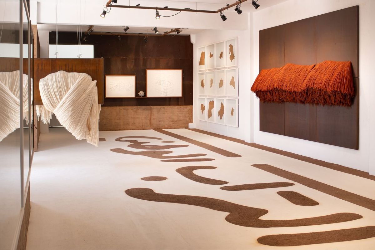 A white and brown art gallery with white and orange textiles on display. Brown Arabic calligraphy is written on the floor. 