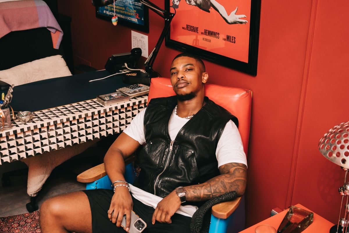 Actor Thabang Molaba seated in orange chair against red wall with classic film posters, wearing white tee and black leather vest in stylish bedroom setting.