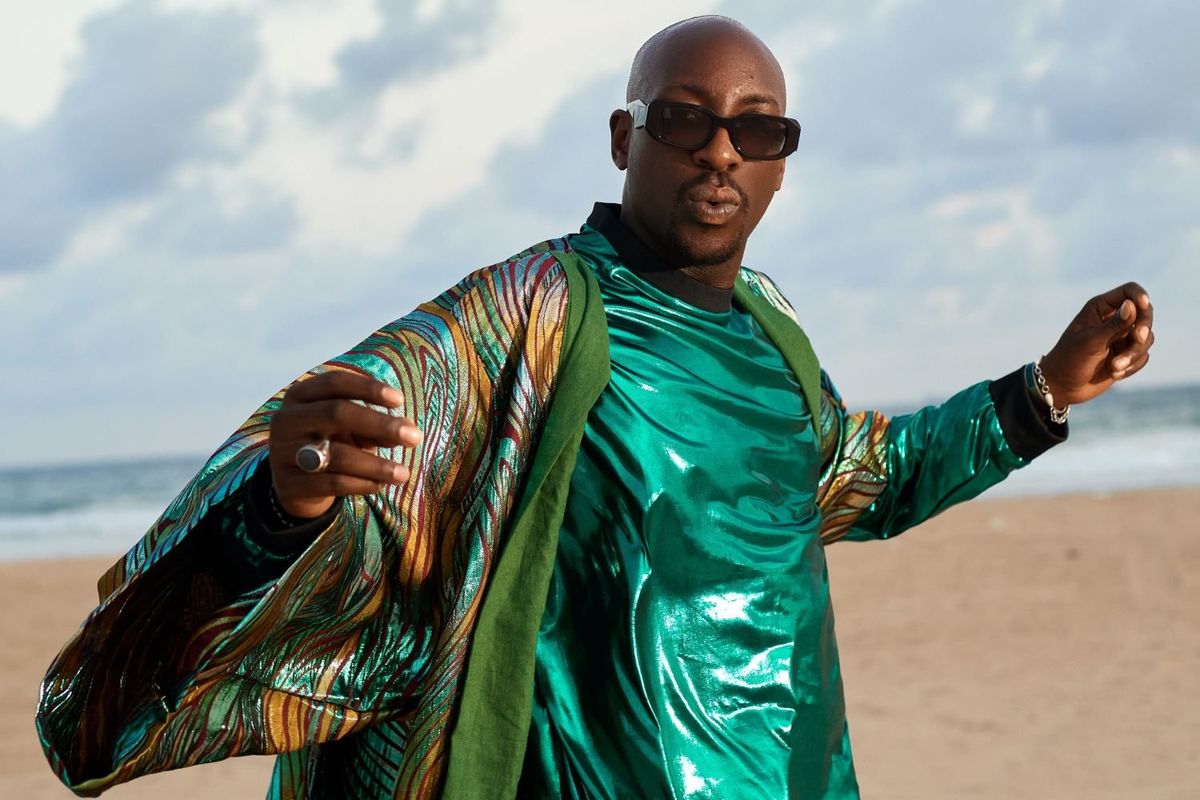 Bien, wearing a shiny green outfit, stands on a sandy beach with the ocean and sky in the background.