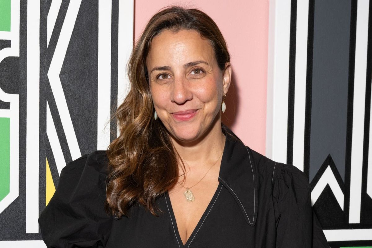 El Glaoui smiling into the camera, wearing a black dress and standing in front of a rose-colored wall adorned with art pieces. 