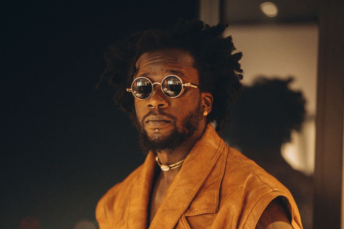 Elijah Kitaka, a Ugandan artist, wearing a sleeveless brown leather vest, round sunglasses, posing with city lights in the background.