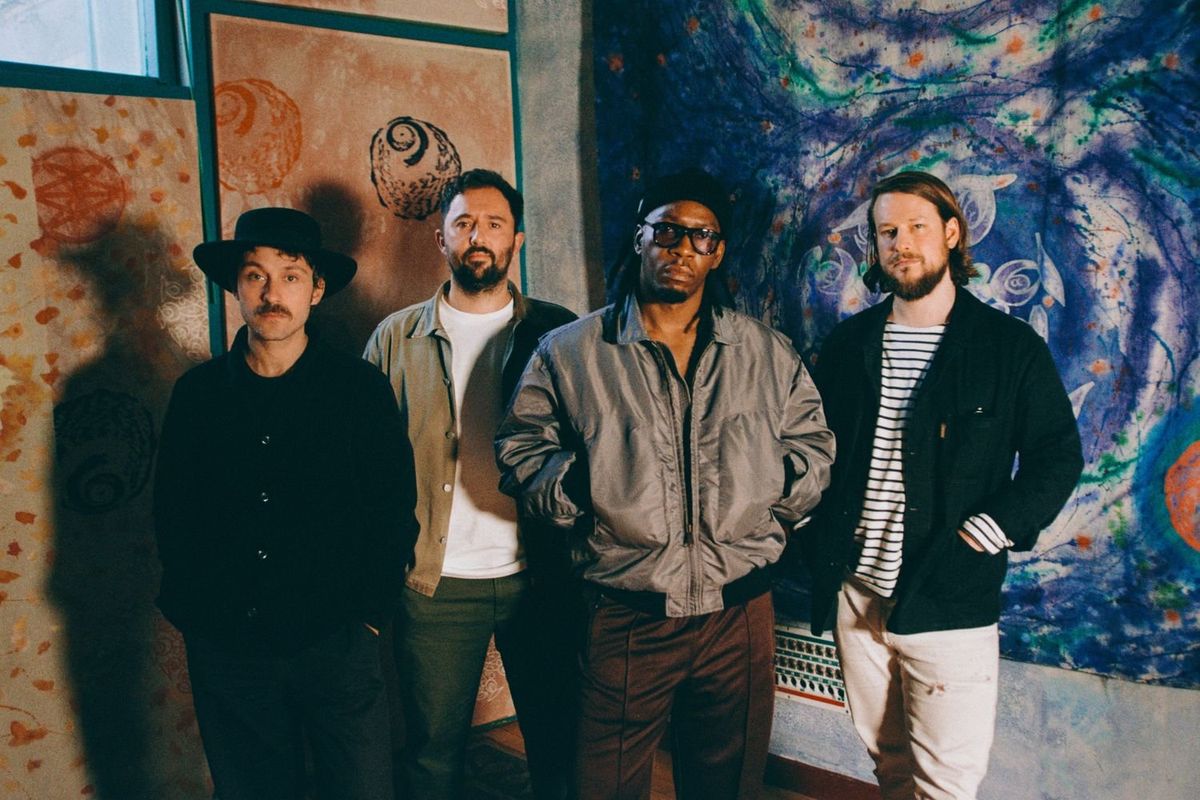 Four men,​ Tiggs Da Author and Thunderdrum, stand side by side in a warmly lit room, each with a distinct style—from casual jackets to a wide-brimmed hat—posed against a backdrop of colorful, abstract art panels.
