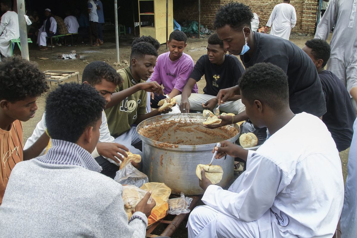 Group of young Sudanese men sharing a communal meal from a large pot, serving food into plastic bags in an outdoor community setting.