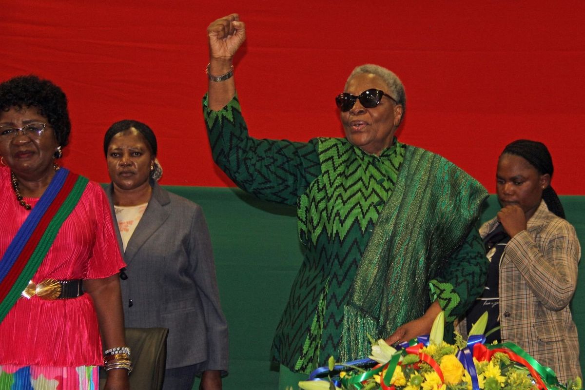 Namibia's first female president, Netumbo Nandi-Ndaitwah stands with her fist in the air. She is smiling and wears a green outfit and sunglasses. There are three women next to hear and a bouquet of flowers in front of her.