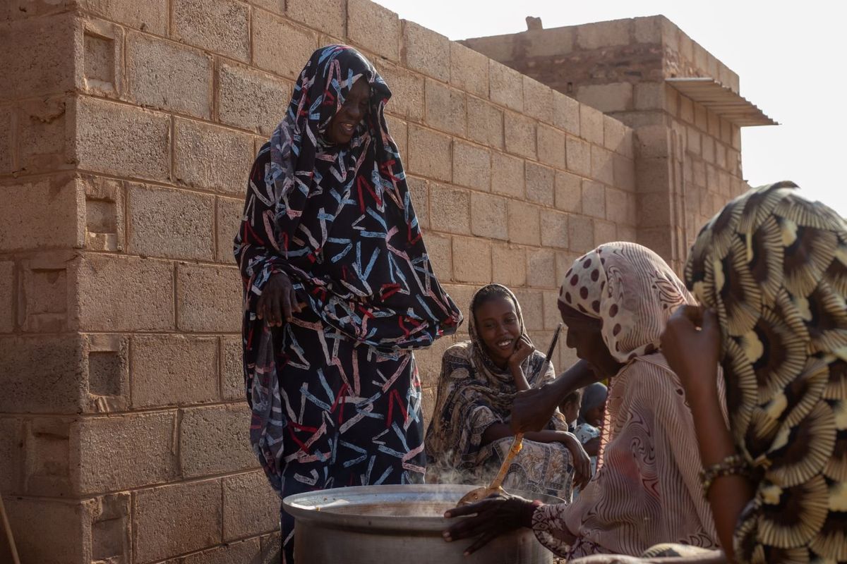 Next to a brick wall, four women wearing toubs with colorful patterns stand or sit around a large round pot, smiling and chatting while one woman stirs the pot's contents with a wooden stick. 