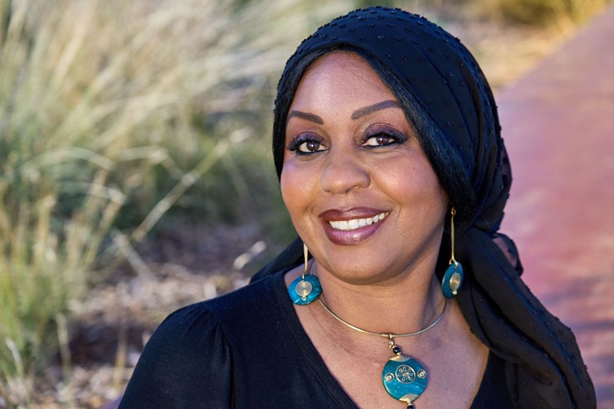 Portrait of Sudanese American radio journalist Hana Baba as she smiles into the camera, wearing a black hijab and blue and golden jewellery. 