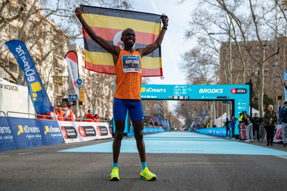 Runner wearing bib number labeled 'KIPLIMO' in orange jersey and blue shorts celebrates victory at the eDreams Mitja Marato de Barcelona half marathon finish line, holding up Uganda's flag. He is standing on a blue carpet at the finish area with race barriers and sponsor banners visible in the background.