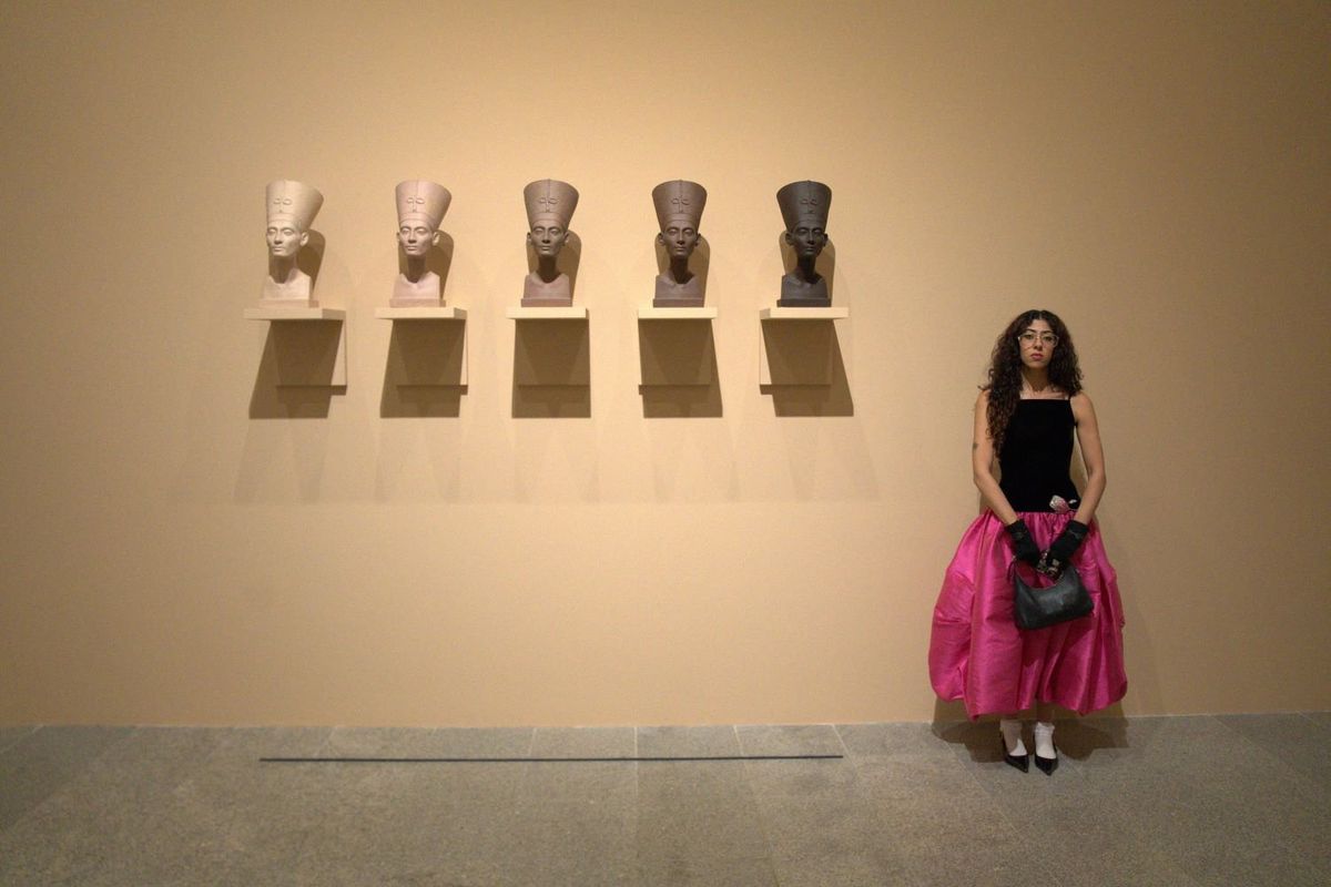 Sara Elmessiry wearing a black shirt and pink skirt, standing in front of five Nefertiti busts in different skin colors ranging from beige to dark brown. 