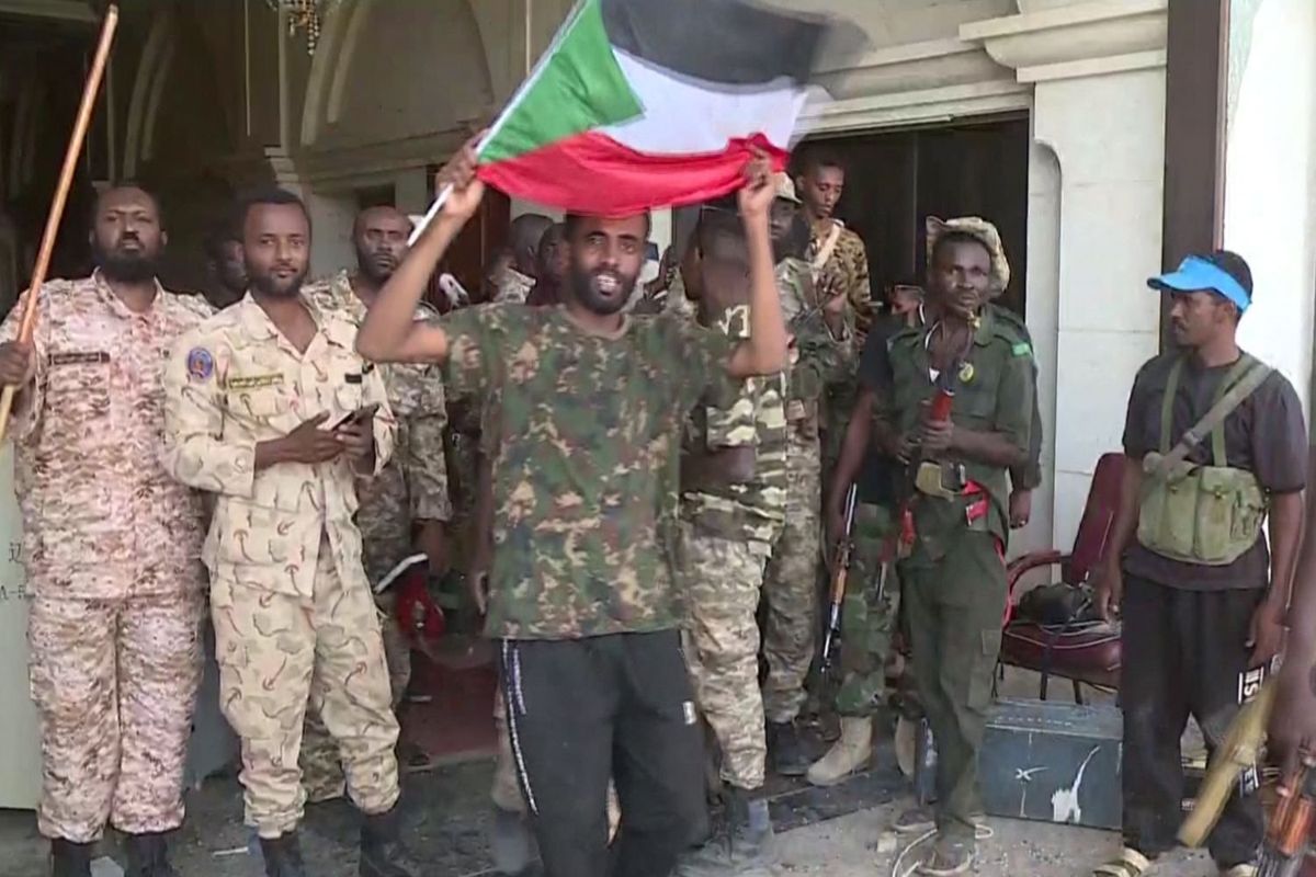 Sudanese army soldiers posing for a picture in the presidential palace in Khartoum. A man in acamouflage T-shirt is standing in front, waving the a Sudanese flag.