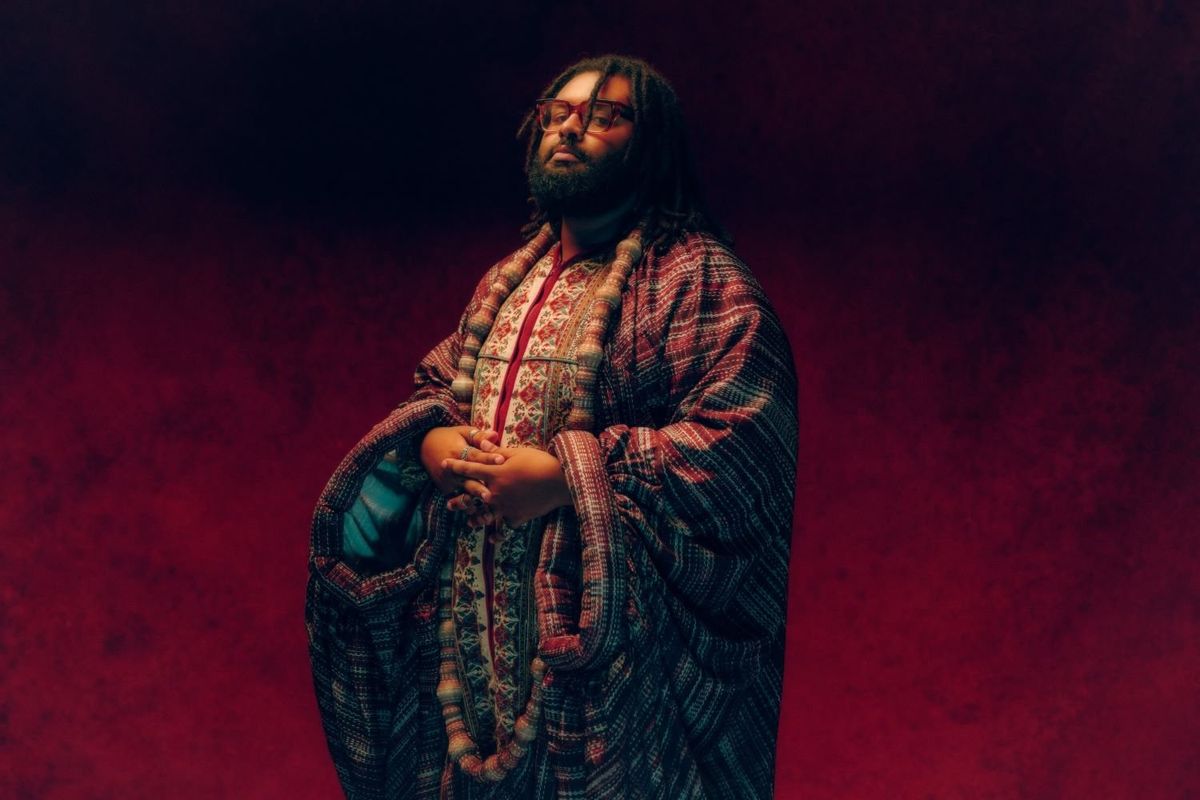 Sudanese British musician Elmiene stands in front of a burgundy background, wearing an elaborate red and white kaftan, silver rings and red glasses. 