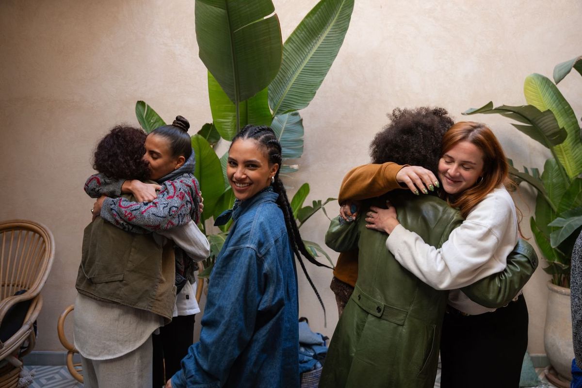 Two pairs of women are hugging in a courtyard with huge banana leaves. In their middle is a young woman with cornrows, wearing a denim shirt, smiling directly into the camera.