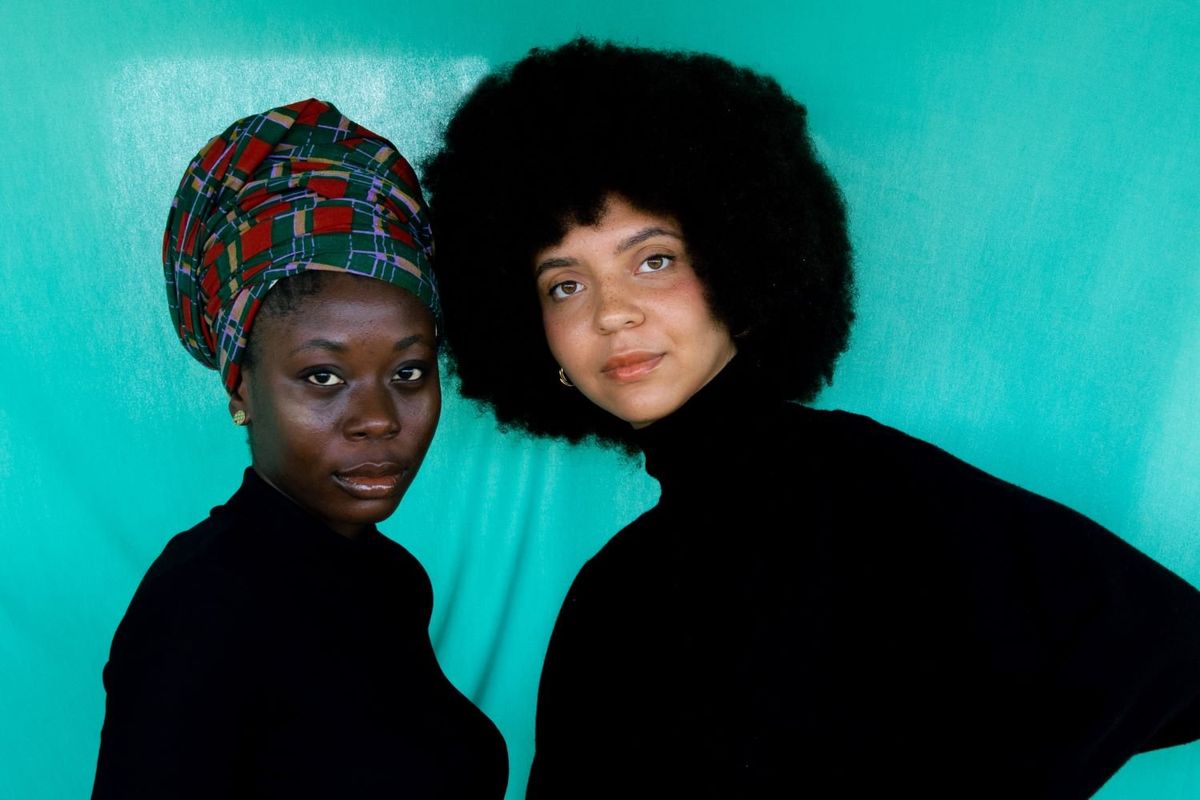 Two women stand leaning head to head with each other and dressed in black.