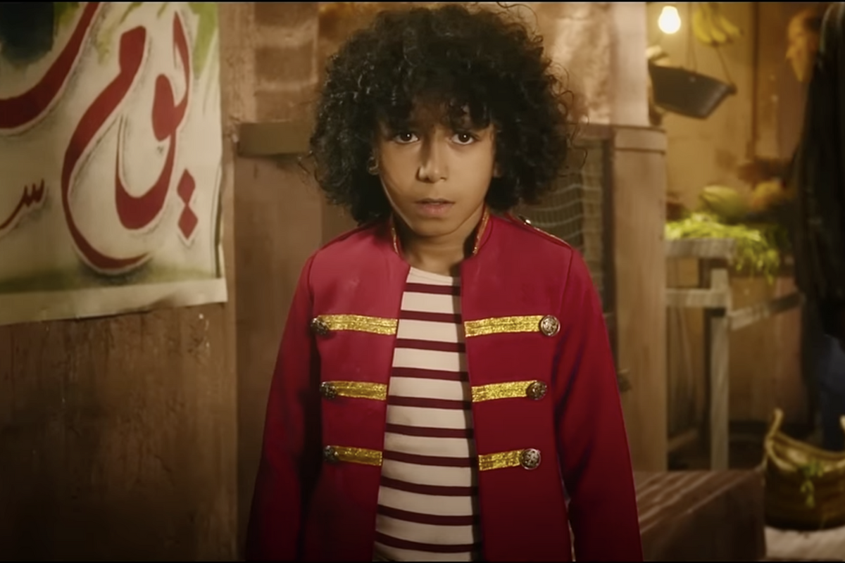 A young Moroccan boy with curly hair, wearing a red vest, looking into the camera.