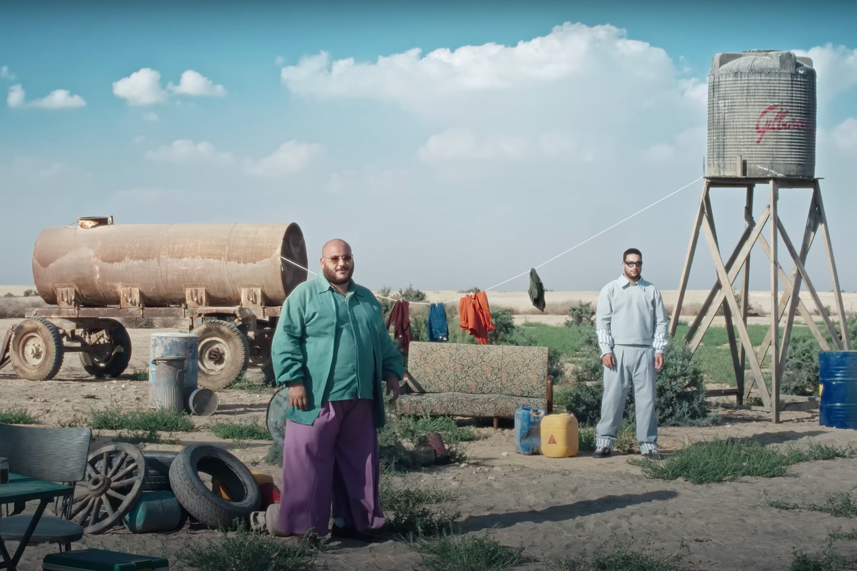 Ahmed Basyoni, wearing purple flowy pants, a turquoise jacket and shirt, is standing on a muddy field with Hussein Gamal, wearing a grey tracksuit.