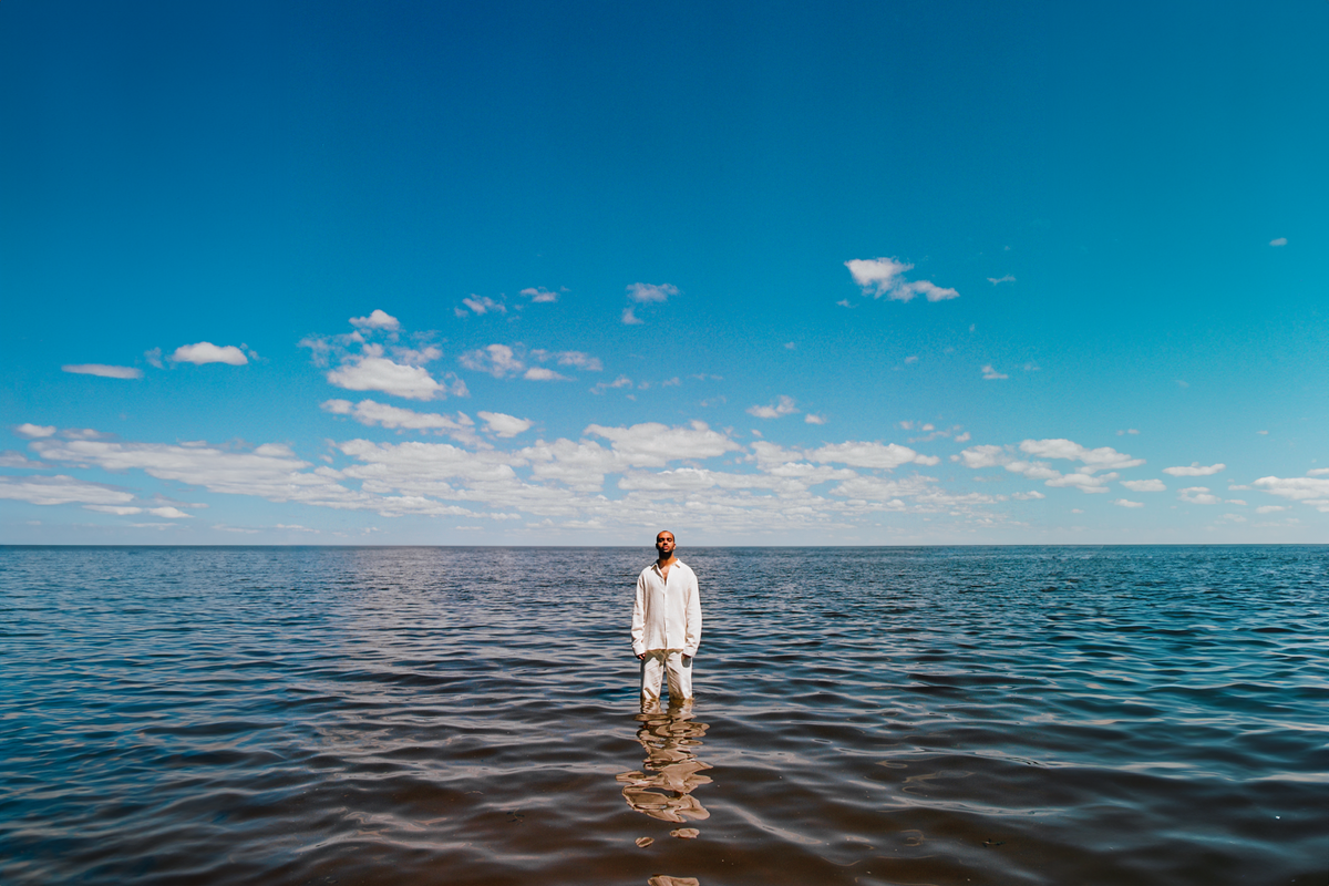 Egyptian French musician Ash standing knee deep in the sea, wearing a white shirt and white pants. 