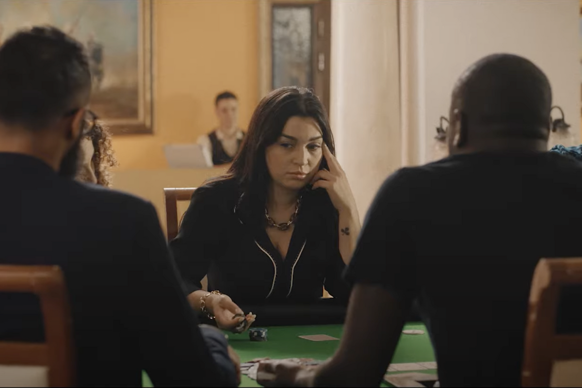  Farah El Fassi, aka Noura, sits at a table gambling with a group of men, wearing a black blouse and appearing deep in thought. 