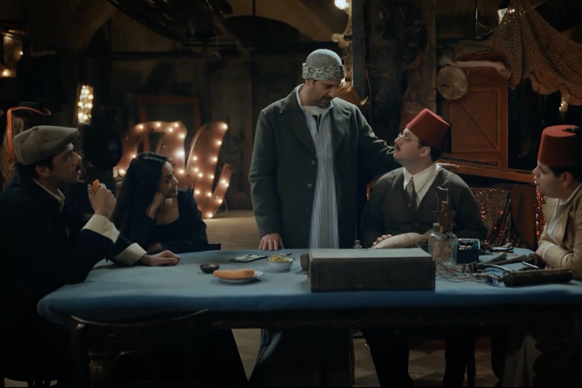 Four men, two wearing the traditional Egyptian red fez hat, and a woman are gathered around a table in a room filled with theater equipment, plotting a scheme. 