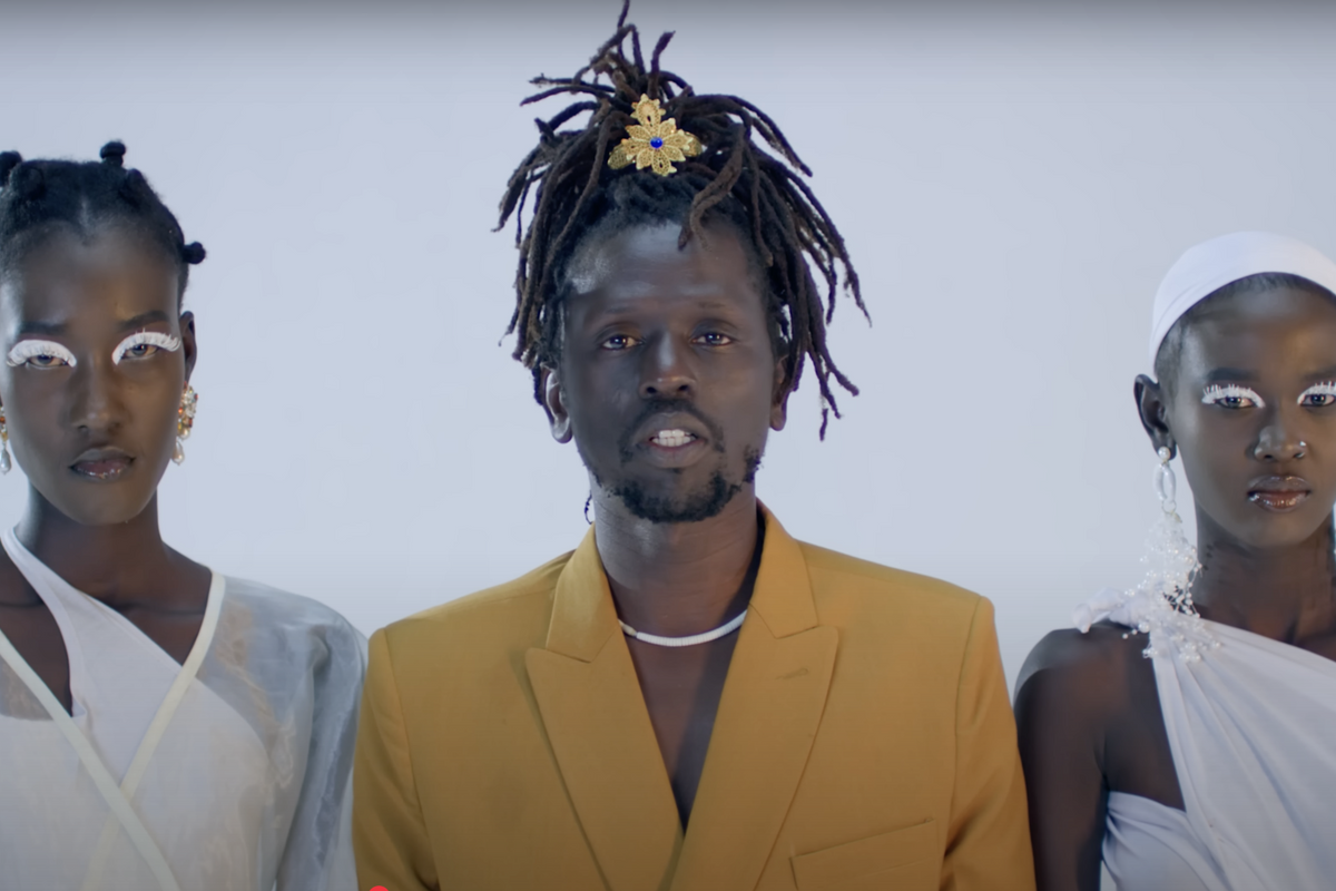 Front view of Emmanuel Jal wearing a yellow suit jacket, a white necklace, and golden flower-shaped broach in his hair. To his left and right are two women wearing all-white dresses, jewelry, and white eyeliner. 