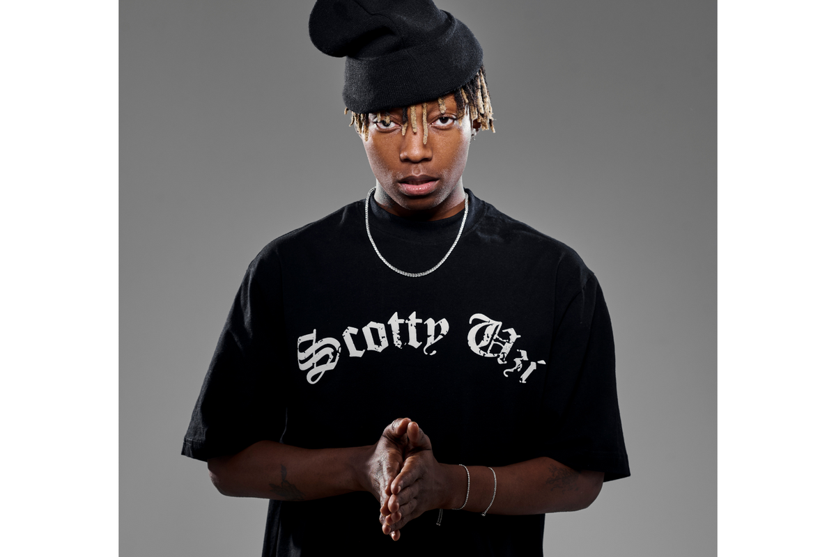 Xduppy stands against a plain gray background, gazing directly into the camera with a serious expression. He wears a black oversized T-shirt that reads “Scotty Uzi” in bold gothic script and a slouchy black beanie tilted to one side, partially covering his bleached-tipped locks. His hands are clasped together in front of him, and he accessorizes with a silver chain necklace and matching bracelets.